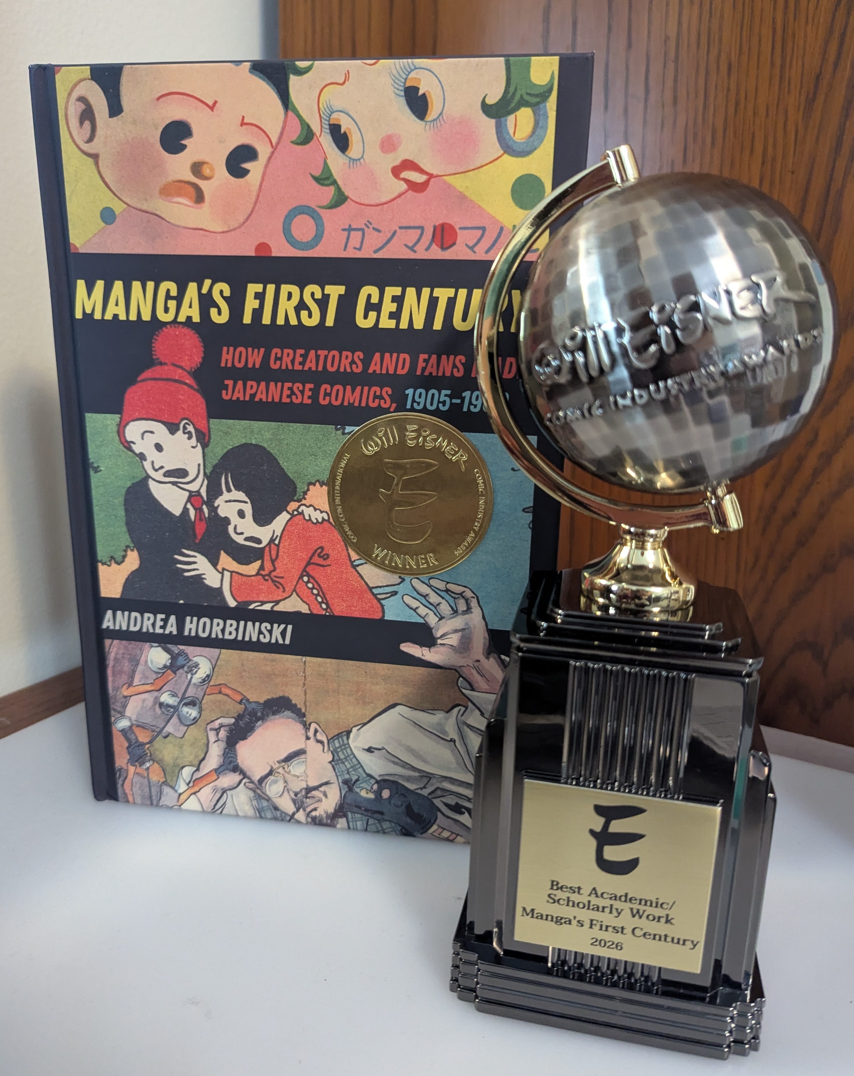 A Will Eisner Award statuette with the plaque bearing the category and work title, placed in front of a copy of Manga's First Century with the gold "Will Eisner Award" winner sticker placed on the cover.